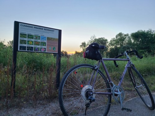A bicycle rests beside a sign for Cumming Prairie Remnant at sunset, with lush greenery in the background.