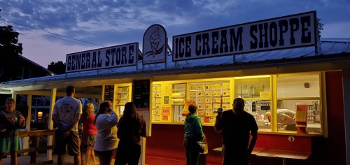 A general store and ice cream shoppe at dusk, with people lined up outside enjoying the evening atmosphere.