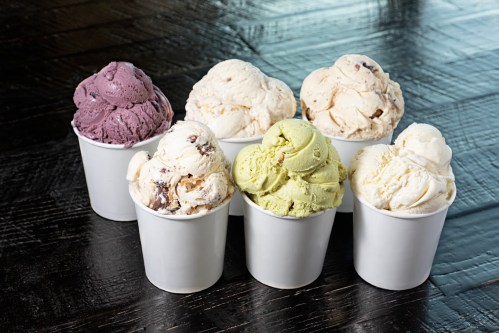 Six cups of ice cream in various flavors: purple, white, and green, arranged on a dark wooden surface.