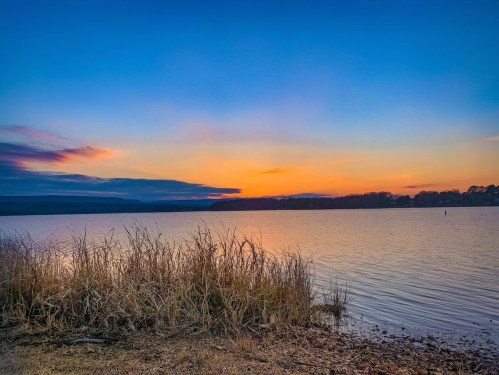 A serene lake at sunset, with vibrant orange and blue skies reflecting on the water and tall grasses in the foreground.