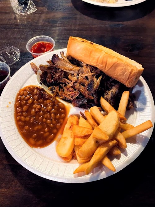 A plate of pulled pork, baked beans, French fries, and a sandwich bun on a paper plate.