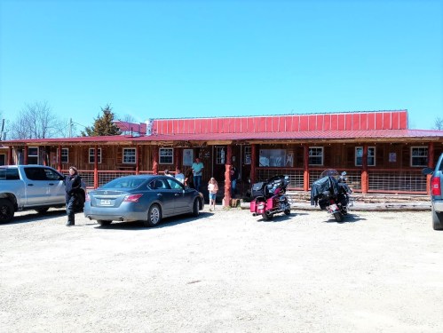 A rustic building with a red roof, surrounded by parked cars and people, on a sunny day.
