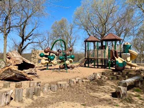 A playground with slides, climbing structures, and swings, surrounded by trees and a sandy area.