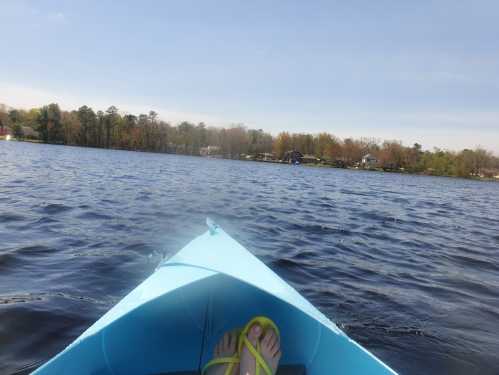 A view from a blue kayak on a lake, with feet in flip-flops visible and trees lining the shore in the background.