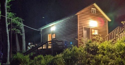 A wooden cabin at night, illuminated by two lights, surrounded by greenery and trees.