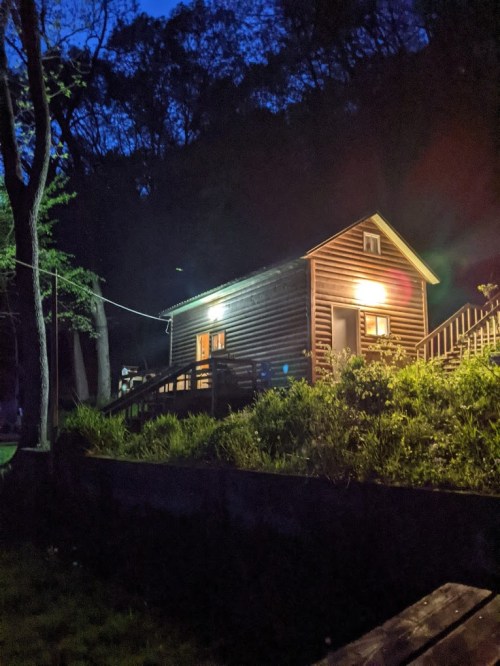 A cozy wooden cabin illuminated at night, surrounded by trees and greenery.