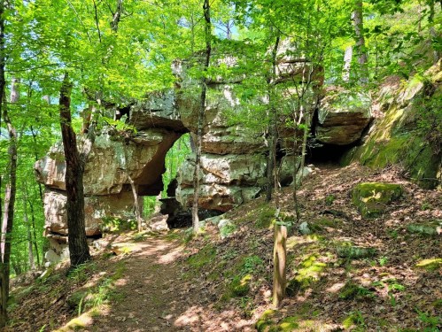 A natural rock arch surrounded by lush green trees and a forested trail.