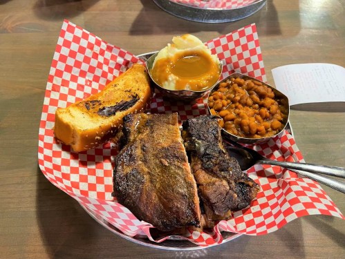 A plate with ribs, mashed potatoes, baked beans, and a slice of garlic bread, served in a checkered basket.
