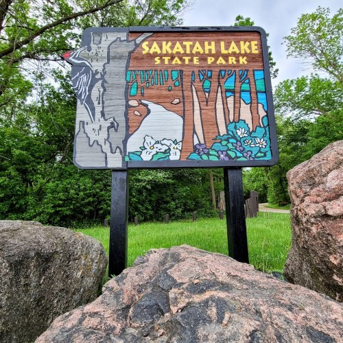 Sign for Sakatah Lake State Park featuring a woodpecker and nature scene, surrounded by greenery and rocks.