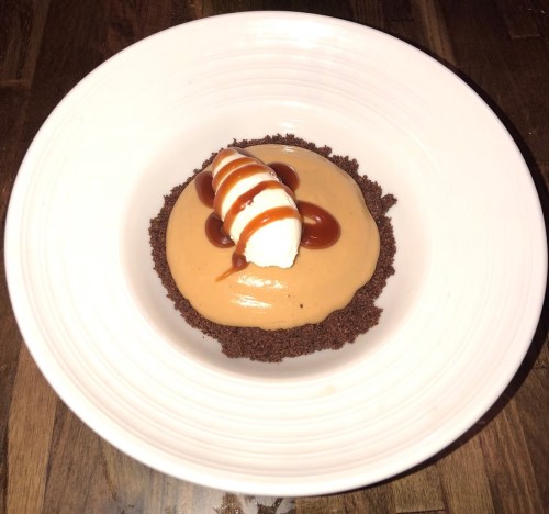 A dessert in a white bowl featuring a chocolate crust, creamy filling, whipped cream, and caramel drizzle.