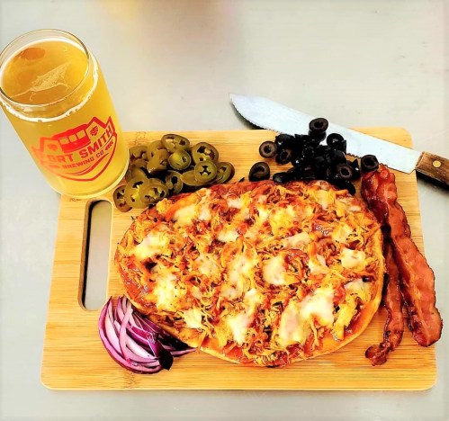 A wooden board with a cheesy pizza topped with pulled pork, bacon, jalapeños, olives, red onion, and a beer.