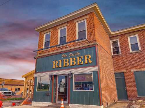 A brick building with "The Scioto Ribber" sign, featuring large windows and a colorful sky in the background.