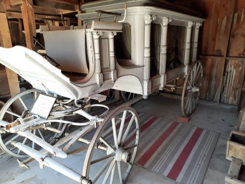 A vintage horse-drawn carriage displayed in a wooden barn, featuring ornate details and a striped rug underneath.