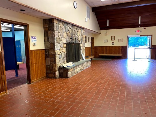 Interior of a room with a stone fireplace, wooden walls, and a doorway leading to a locker area. Natural light from the entrance.