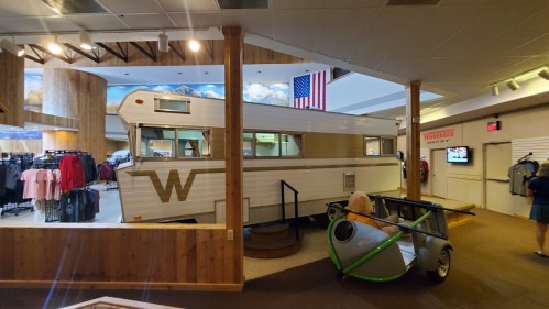 A vintage camper displayed indoors, with an American flag in the background and clothing racks nearby.