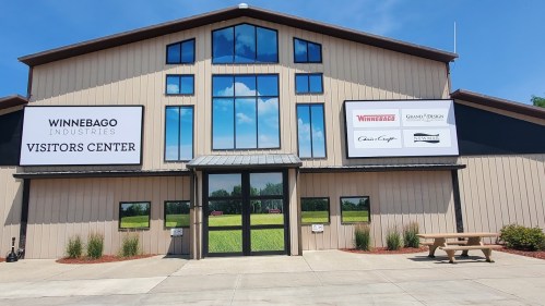 Exterior of the Winnebago Industries Visitors Center, featuring large windows and signage, with a clear blue sky above.
