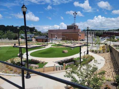 A sunny park with green lawns, pathways, and seating areas, surrounded by buildings and a blue sky with clouds.