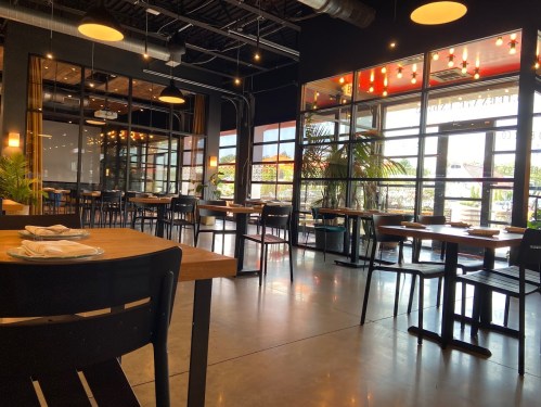 Modern restaurant interior with wooden tables, black chairs, large windows, and plants, creating a bright atmosphere.