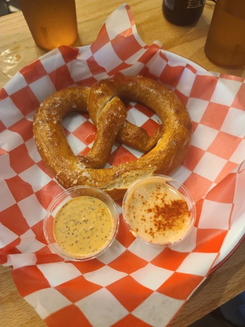 A large, soft pretzel on a red and white checkered paper, accompanied by two dipping sauces in small cups.