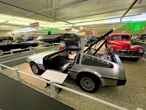A DeLorean car with its gullwing doors open, displayed in a vintage car museum alongside other classic vehicles.