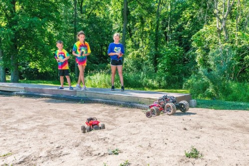 Children in colorful shirts control remote-controlled cars on a dirt track in a sunny park.