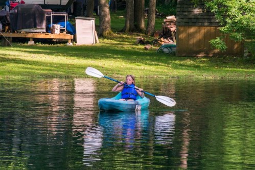 A girl paddles a kayak on a calm lake surrounded by trees and a cabin in the background.