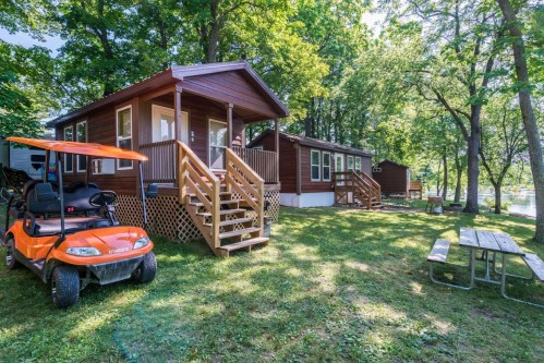 A cozy cabin surrounded by trees, with a golf cart parked nearby and picnic tables on the grassy lawn.