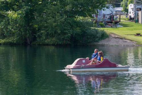 Two people enjoy a pedal boat ride on a calm lake surrounded by greenery and recreational vehicles.