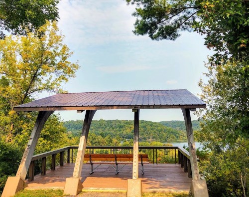 A wooden pavilion with a slanted roof overlooks a serene lake surrounded by lush green trees.