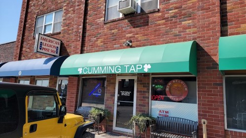 A brick building with a green awning reading "Cumming Tap" and an "ATM Inside" sign, with benches outside.