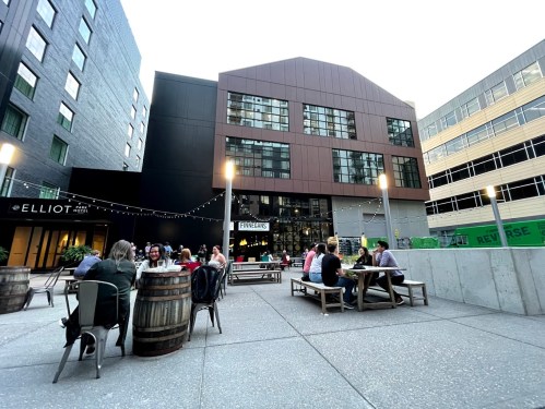 Outdoor dining area with people at tables, string lights, and modern buildings in the background.