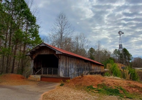 A rustic wooden covered bridge beside a dirt path, with trees and a windmill in the background under a cloudy sky.