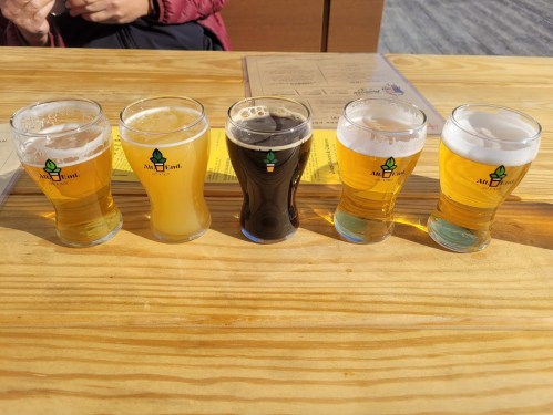 Five small glasses of beer in a row on a wooden table, featuring different colors and styles.