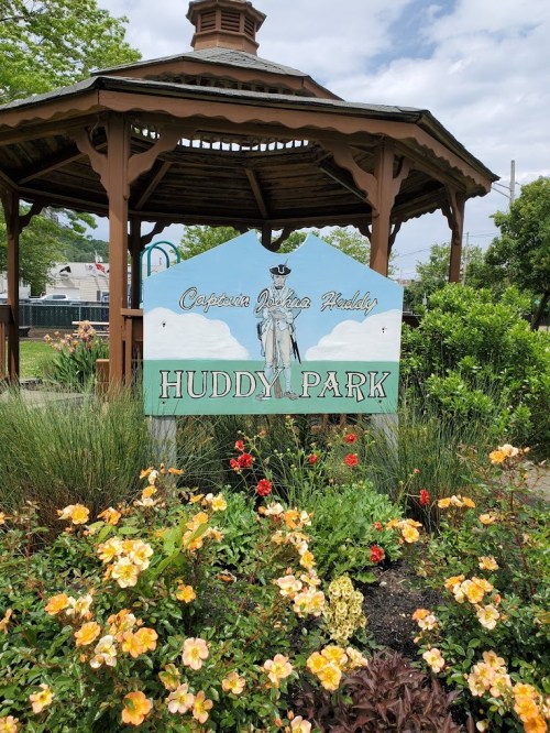 Sign for Huddy Park with a statue and flowers in the foreground, set against a cloudy sky.