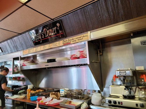 Interior of Rosy's Diner featuring a counter, kitchen area, and menu sign. A staff member is preparing food.