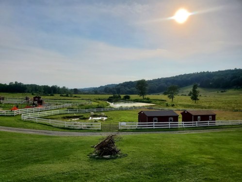 A serene farm landscape with a pond, red barns, and a bright sun overhead, surrounded by green fields and hills.