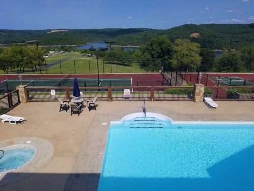 A scenic view of a pool area with tennis courts and a lake in the background, surrounded by green hills.