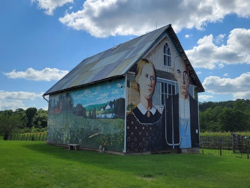 A barn with a large mural depicting two figures against a scenic background, under a blue sky with clouds.