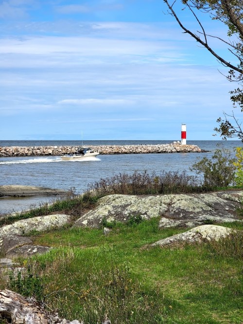 A boat navigates near a rocky shore with a red and white lighthouse in the background under a blue sky.