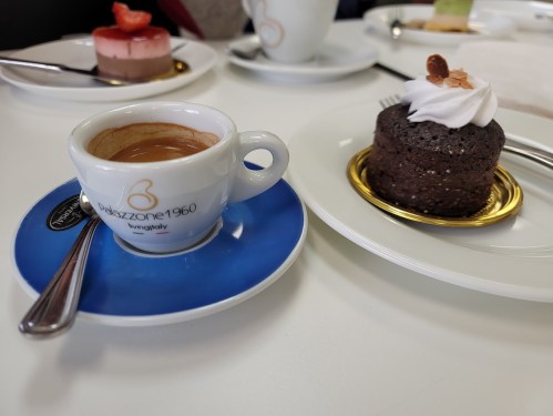 A small cup of espresso on a blue saucer next to a chocolate dessert topped with cream and a nut.