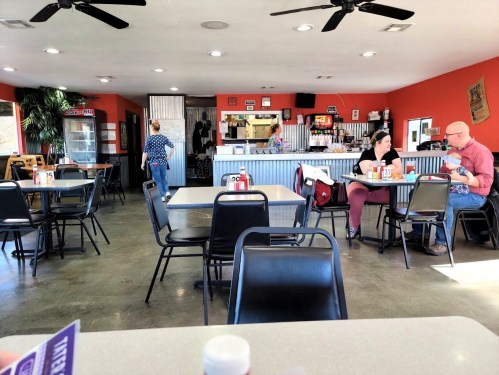 A casual diner interior with empty tables, patrons eating, and a counter in the background. Brightly colored walls.