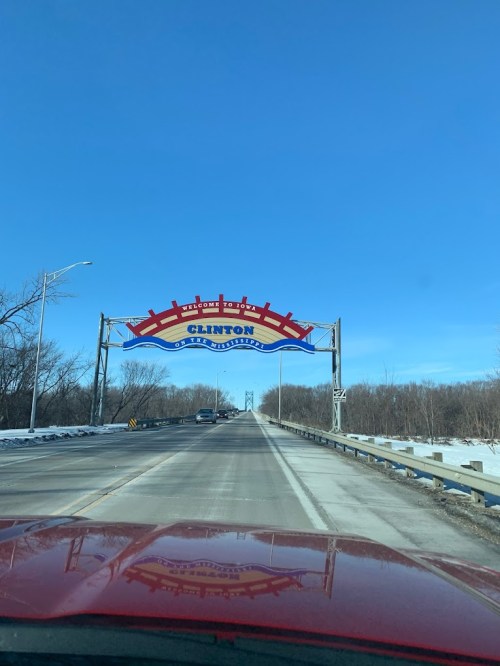 Welcome sign for Clinton, with a clear blue sky and a snow-covered road ahead.