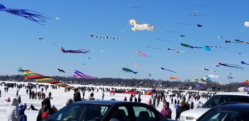 A snowy landscape filled with people flying colorful kites, with various kites soaring in the blue sky above.