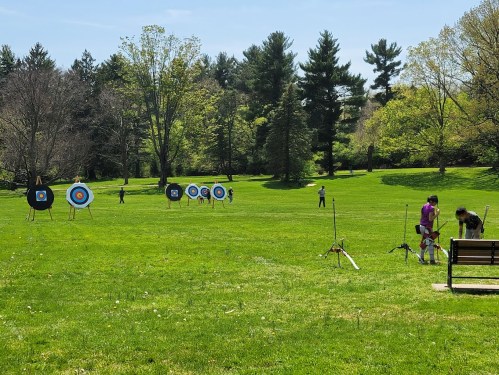 A sunny park scene with archery targets set up on a grassy field, people practicing archery in the background.