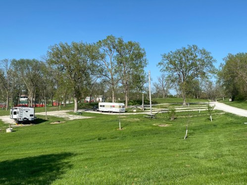 A grassy area with trees, a gravel road, and parked trailers under a clear blue sky.