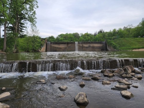 A serene view of a waterfall cascading over a dam, surrounded by lush greenery and rocky riverbank.