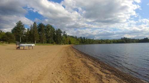 A sandy beach curves along a calm lake, surrounded by trees under a partly cloudy sky.