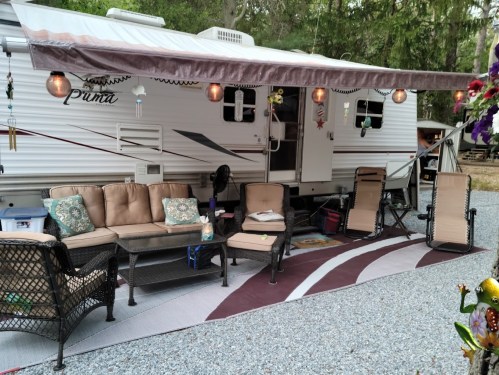 A cozy outdoor setup with a camper, seating area, and decorative lights on a gravel surface surrounded by trees.