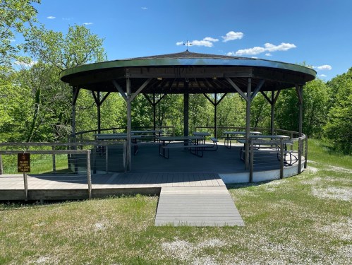 A circular wooden gazebo with a metal roof, surrounded by greenery and a gravel path on a sunny day.