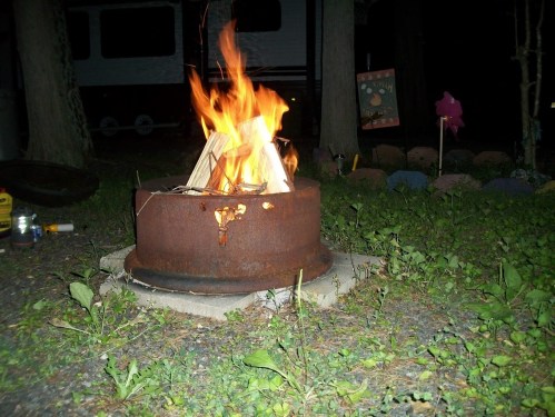 A campfire burns brightly in a metal fire pit, surrounded by grass and trees at night.